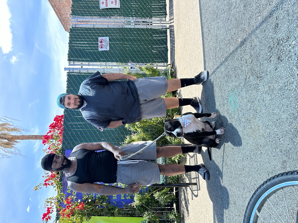 Two Skid Row neighbors posing with their pit bull puppy in a fresh white bandana after a full spa day inside the mobile van